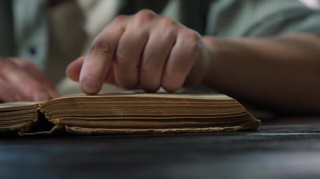 Woman reads book while sitting at table in dimly lit room, her finger tracing text on page close-up