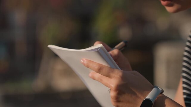 White woman writing in notebook outdoors, closeup of hands holding pen and paper, striped shirt, smartwatch and ring, warm sunset light, blurred urban rooftop background, candid reflective mood