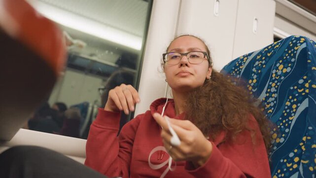 Young latina woman on train window, leaning back with earbuds and red hoodie, soft cabin light, reflective gaze toward passing city, quiet night journey capturing candid urban solitude and tired rest