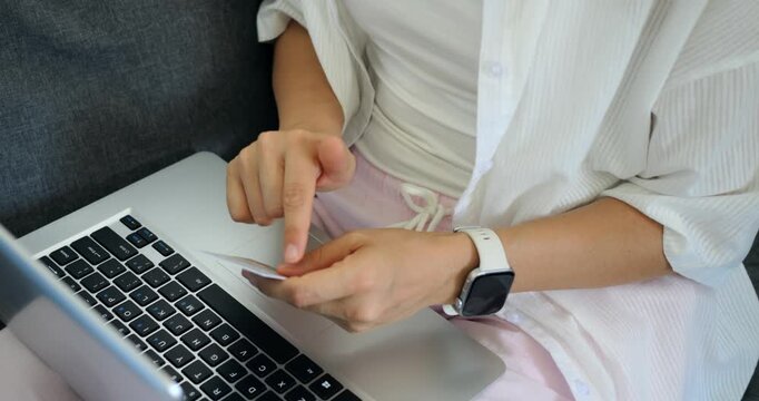 Woman holding smartphone and typing on laptop performing verification or multitasking between devices, closeup hands and smartwatch, peaceful living room environment and focused task