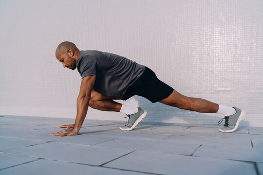 Side view of athlete doing deep lunge stretch on outdoor floor, morning fitness and mobility workout, urban training environment, healthy man exercising in sunlight, strength and balance concept.