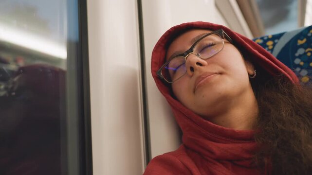 Relaxed sleeper leaning on train window, red hoodie, glasses, night blur outside, reclining seat, soft overhead light, long hair, serene expression, weary commuter finding respite