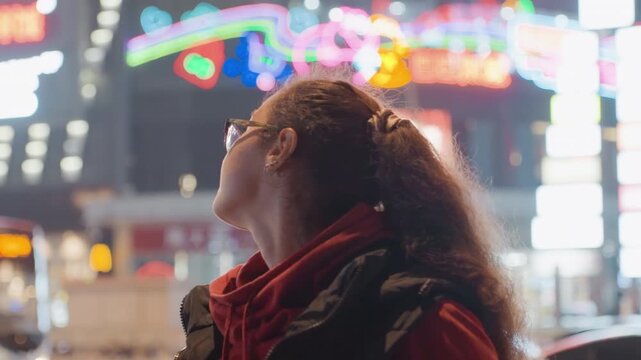 Young woman under neon signage night gesturing to passersby pointing toward transit stop expressive hand movements streetwear hoodie backpack candid urban interaction vibrant glow