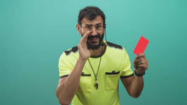 Man referee in neon shirt holds red card and whistle, hand cupped to mouth as if shouting, headset mic and beard visible in studio setting; discipline stern.