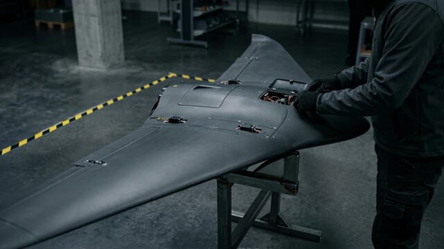 A person works on assembling a military drone in a dimly lit workshop. The individual inspects the drone's components and prepares it for a test flight outside