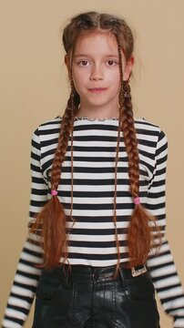 Cheerful funny young school girl showing tongue making faces at camera, fooling around, joking, aping with silly face teasing bullying, abuse. Preteen female child kid 10 years old on beige background
