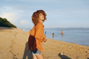 A young woman with curly hair wearing an orange shirt and denim shorts stands on a sandy beach...