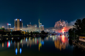 Fototapeta premium Fireworks display over Main River waterfront at night in Gutleutviertel, Frankfurt am Main, Hessen, Germany with illuminated skyline reflections