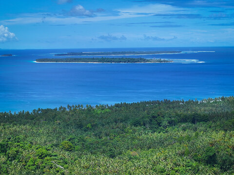AERIAL: View across lush tropical forest toward long low islands surrounded by deep blue ocean and pale reef lines. A striking island seascape from the Mentawai archipelago in West Sumatra, Indonesia.