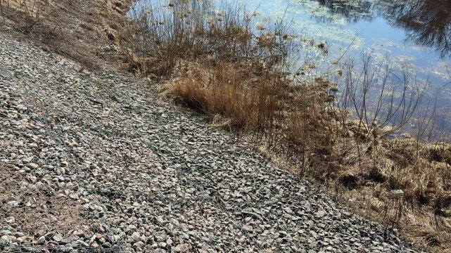 Crushed stone riverbank with dry grass, stainless wire mesh over pebble embankment beside calm river, textured closeup of rocks.