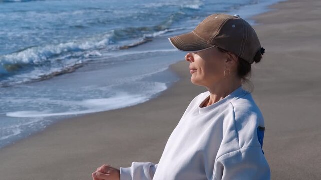 Mature woman enjoying the sunshine on a sandy beach. Serene middle-aged woman in a baseball cap relaxing on a beach chair, thoughtfully watching the gentle ocean waves on a sunny day