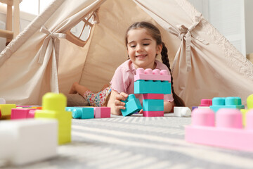 Little girl playing with building blocks near toy wigwam in playroom, low angle view © New Africa