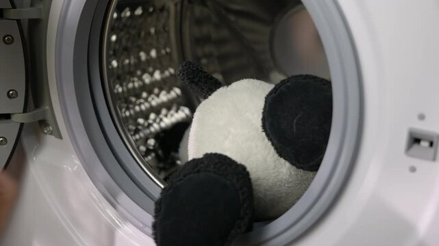 Person loading teddy bear and plush toys into a washing machine. Careful hands placing several plush toys, including a teddy bear and a panda, into the drum of a modern washing machine