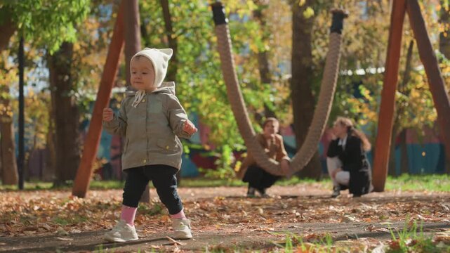 Young child walks confidently by colorful playground. Little one enjoying outdoor play as family watches happily. Toddler confidently steps near swing amid bright fall sunshine with family support