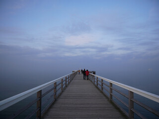 Fototapeta premium A wooden pier extends out over calm water towards a hazy horizon. Silhouetted figures walk along the pier under a pale, cloudy sky.