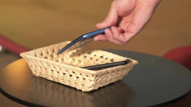 Person placing smartphones and headphones in a basket. Person placing personal items like two smartphones and a pair of headphones into a small wicker basket sitting on a black table