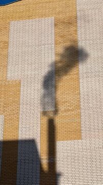 Shadow gas pipe smoke on brick facade under clear blue sky, sunlight casts dark silhouette and elongated pipe shadow.