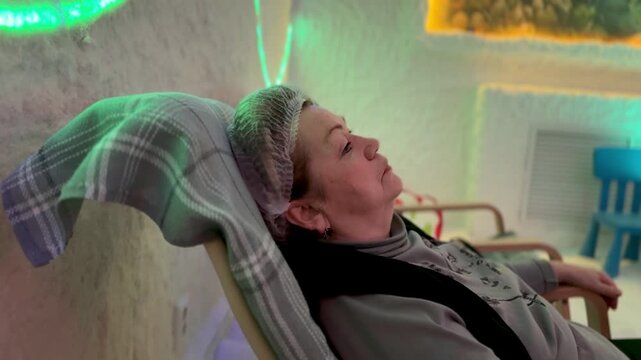 Elderly woman reclining in salt cave halotherapy chamber wearing cap on recliner under halogen neon lighting salt encrusted walls.