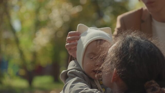 Closeup baby hat adjustment by grandmother in sunny park with father watching, gentle touch and knit cap detail, tender caregiving exchange highlighting family warmth and intergenerational bond