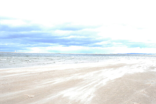 A wide, sandy beach is covered in blowing sand under a cloudy sky. The ocean stretches out in the distance, with a faint horizon line.