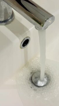 Dirty stainless faucet pouring water into unclean acrylic sink, visible mineral stains and soap scum around drain, slow steady.