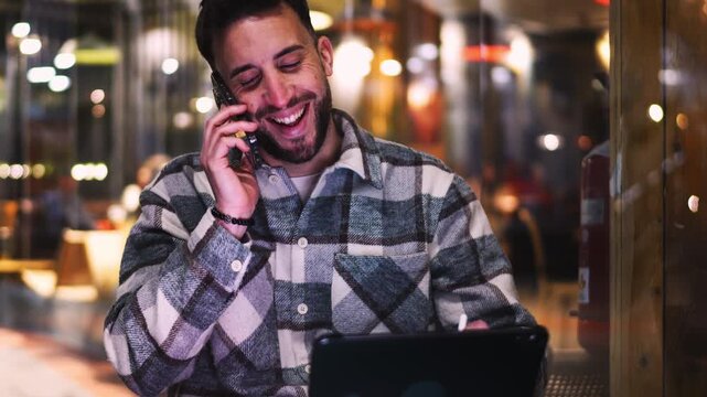 Happy man talking on a smartphone while multitasking with a digital tablet in a cozy interior, expressing joy, success and professional confidence in a modern work environment
