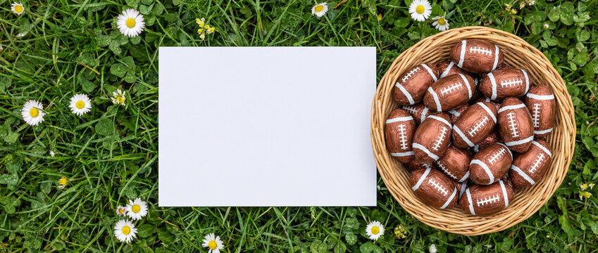  Blank white greeting card mockup and basket of American football chocolate Easter eggs on spring green grass with daisies. Flat lay top view