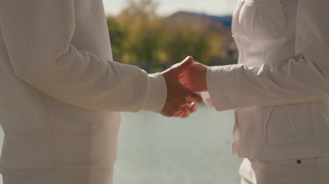 Lakeside vow exchange between caucasian partners. Two women in white sweaters hold hands by tranquil lake, soft autumn light, gentle touch and quiet smiles as they commit to marriage and future family