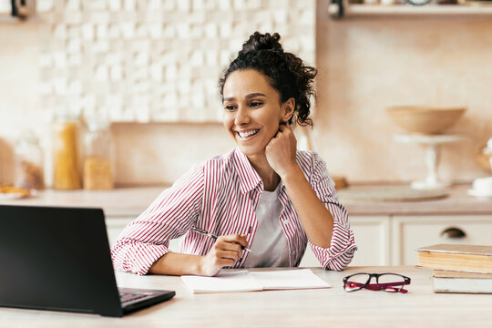 A woman sits at a wooden table with a laptop in front of her. She smiles while looking at the screen. A notebook and glasses are placed next to her. The setting is bright and casual.