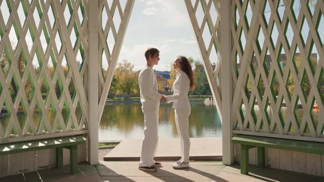 Couple exchanging bouquet by lake, pavilion frames scene, hands meet holding flowers, gentle breeze, smiles and eye contact, sunlight reflecting on water, ceremony vibe and family promise undertones