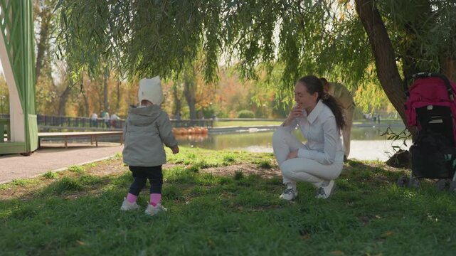 White mother kneeling by lakeside tree, toddler approaching stroller. Gentle encouragement under willow shade, sunlight on grass, supportive parenting moment, clapping and smiles, teaching walking