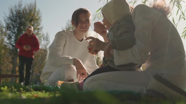 Close white family feeding toddler outdoors, mother offers snack while father watches with smile, candid park scene with passerby in background, sunlit grass and playful laughter, caring married