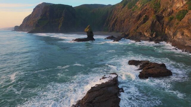 Waves crash against the rugged cliffs and sea stacks of Karekare Beach, New Zealand. The wild, untamed beauty of the coastline is a popular destination for surfers.
