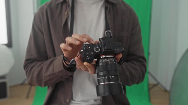 Man holding dslr camera and turning top dial with fingertips in studio with green screen and softbox lights visible; focus calm.