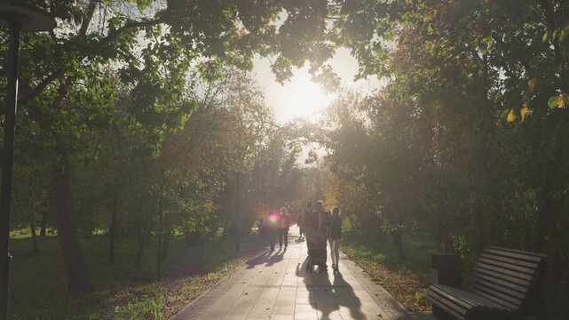 Stroller family walking down sunlit path in leafy park, parents pushing baby carriage between benches and trees, long shadows and autumn leaves, relaxed weekend stroll capturing parenting and married