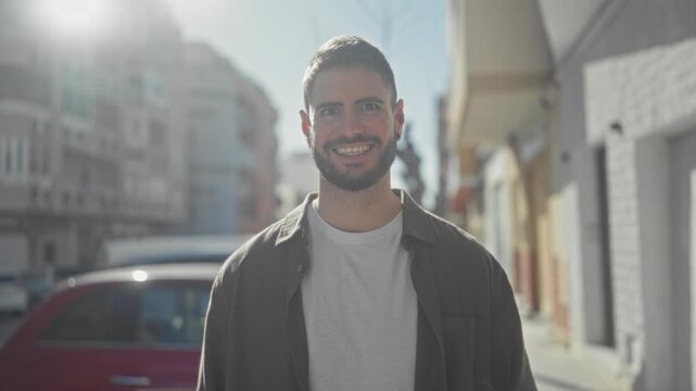 Young hispanic man smiling with visible face and beard, wearing white tshirt and open shirt while standing on a sunny street; optimism approachable warmth.