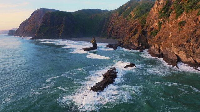 Waves crash against the rugged cliffs and sea stacks of Karekare Beach, New Zealand. The wild, untamed beauty of the coastline is a popular destination for surfers.
