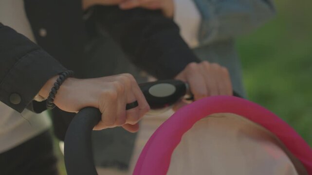 Parenting teamwork scene. Child stroller with pink canopy. Closeup of parent holding stroller handle in park. Parents coordinating to push stroller with safety grip amidst park pathway