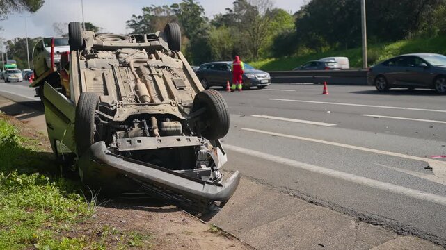 Car lying upside down after an accident on the highway. Policeman directing traffic and allowing other vehicles to pass. Focus on car crash and vehicle insurance