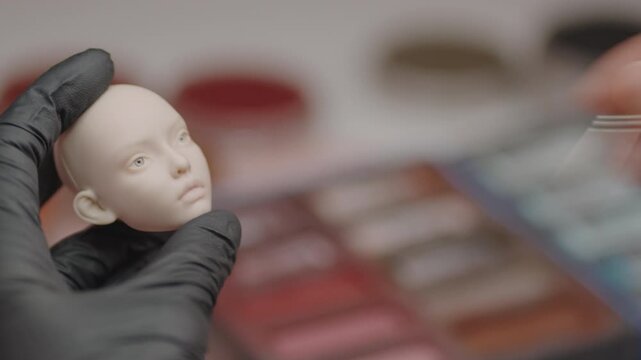 Closeup of skilled artist carefully painting facial details on handmade doll head using fine brush, focusing on craftsmanship and precision