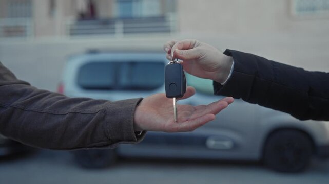 Young hispanic man receives car keys with open palm while another hand drops the fob beside a parked car on street; trust transfer.