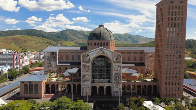 Aerial frontal view of National Shrine basilica facade with dome, tower and religious murals with city and hills in Aparecida, Brazil. Pull back camera movement