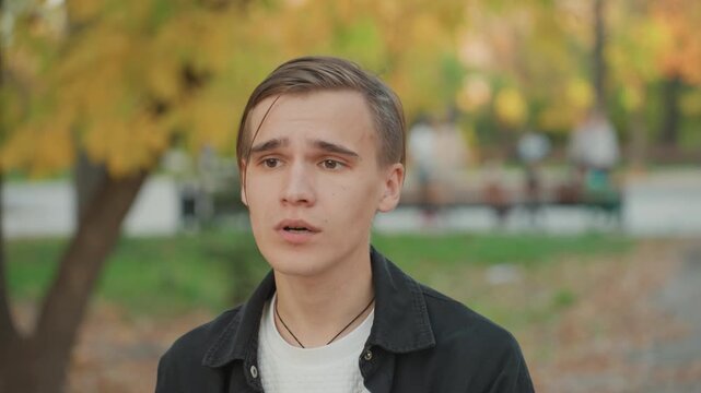 Young caucasian man rehearsing marriage vows in quiet autumn park, closeup portrait showing vulnerable expression, speaking to unseen partner, gentle leaves, bench, jacket and necklace details,
