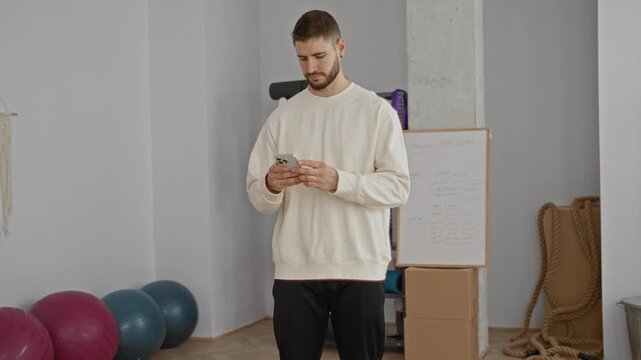 Young man holding smartphone and glancing at the screen in a yoga studio with fitness balls and ropes, checking a routine on his device; relaxed wellness.