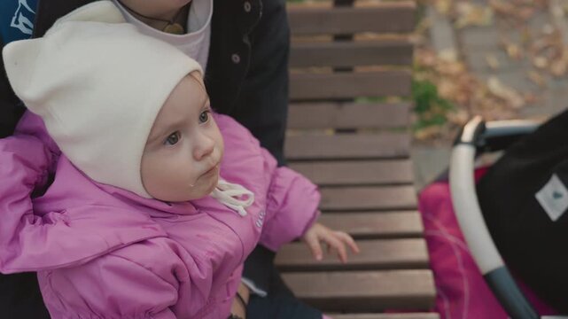 Family baby reaches hand in park. Father holds child on wooden bench as caregiver leans in for gentle touch, stroller at side, autumn leaves on ground, curious gaze and soft kiss on head, intimate