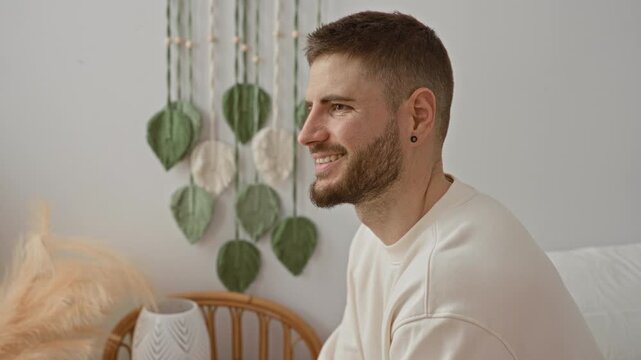 Man face smiling on bedroom bed with macrame wall hanging and pampas grass wearing cream sweater and earring; contentment.