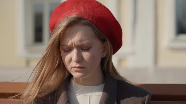 White woman on bench receiving call, red beret fluttering, furrowed brow and worried expression, sunlight highlights hair, urban facade background, moment of decision and resolve, closeup portrait