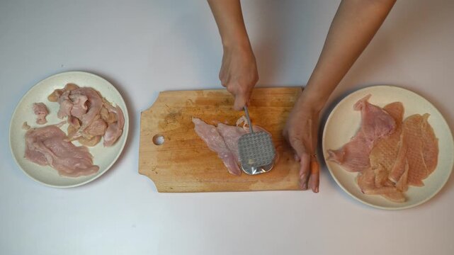 Woman hands placing raw chicken breast on cutting board and pounding with meat hammer