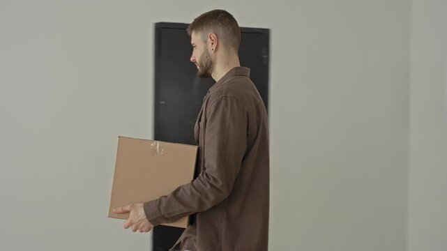 Man lifting a cardboard box with hands, bending at the knees and gripping the parcel inside a bright empty building room with window light; calm transition.