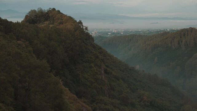 Cinematic misty forest mountain landscape during early morning with soft fog covering the valley. Filmed at Tebing Keraton, Bandung, West Java, Indonesia. Peaceful tropical scenery with lush greenery 